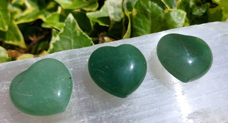 Pu&ograve; includere: Tre pietre a forma di cuore verde lucido. Le pietre sono disposte su una superficie bianca, con fogliame verde sullo sfondo. I cuori hanno una finitura liscia e lucida e variano leggermente nella forma.