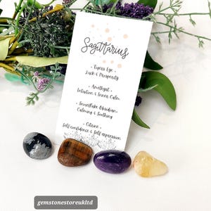 May include: A collection of polished gemstones for the Sagittarius zodiac sign. The image includes a card with the word "Sagittarius" and the gemstones: Tigers Eye, Amethyst, Snowflake Obsidian, and Citrine. The gemstones are in shades of black, brown, purple, and yellow.