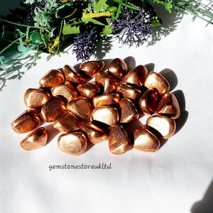 May include: A collection of tumbled copper stones. The stones are a shiny, metallic copper colour and have a smooth, rounded shape.