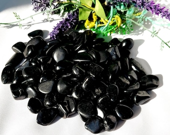 Op de afbeelding: Een stapel gepolijste zwarte obsidiaanstenen. De stenen zijn glad en reflecterend, met een glanzende afwerking. Groen en paars bloemstuk op de achtergrond. De tekst "gemstonestoreukltd" staat onderaan.