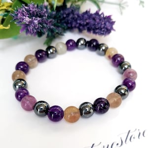 May include: A bracelet made of mixed gemstones in shades of purple, brown, and grey. The bracelet features round beads of varying sizes and colours, including amethyst and hematite. The bracelet is displayed on a white surface.