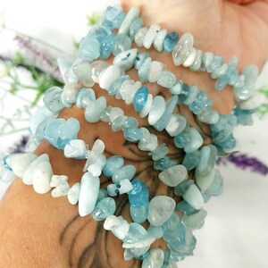 K&ouml;nnte beinhalten: Mehrere Armb&auml;nder aus Aquamarin-Chip-Perlen sind an einem Handgelenk gestapelt. Die Armb&auml;nder bestehen aus hellblauen und wei&szlig;en unregelm&auml;&szlig;ig geformten Perlen. Der Hintergrund ist wei&szlig; mit einem Hauch von lila Blumen.