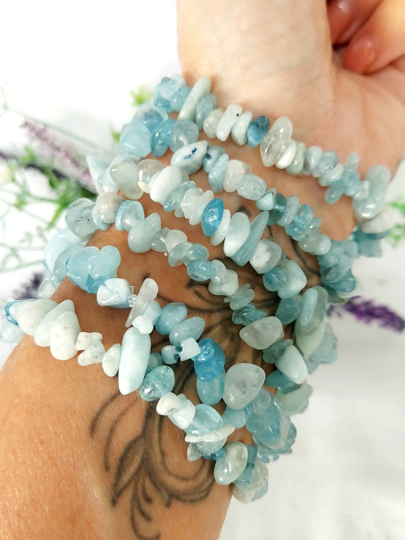 K&ouml;nnte beinhalten: Mehrere Armb&auml;nder aus Aquamarin-Chip-Perlen sind an einem Handgelenk gestapelt. Die Armb&auml;nder bestehen aus hellblauen und wei&szlig;en unregelm&auml;&szlig;ig geformten Perlen. Der Hintergrund ist wei&szlig; mit einem Hauch von lila Blumen.