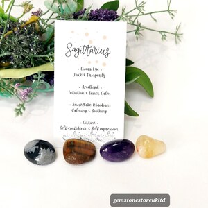 May include: A collection of four polished gemstones: snowflake obsidian, tiger's eye, amethyst, and citrine. A card reads "Sagittarius" with associated properties. The gemstones are arranged on a white surface with floral accents.