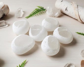 May include: Six white, smooth, carved stone candle holders. The holders are oval-shaped with a hollowed-out center. The image also includes clear quartz crystals, birch bark, and green fern leaves, creating a natural, earthy aesthetic.