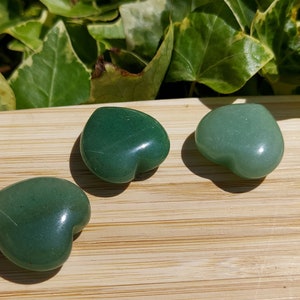 Pu&ograve; includere: Tre pietre di avventurina verdi a forma di cuore su una superficie di legno. Le pietre variano in tonalit&agrave;, da un verde foresta intenso a un verde pi&ugrave; chiaro e pastello. Lo sfondo presenta un fogliame verde.