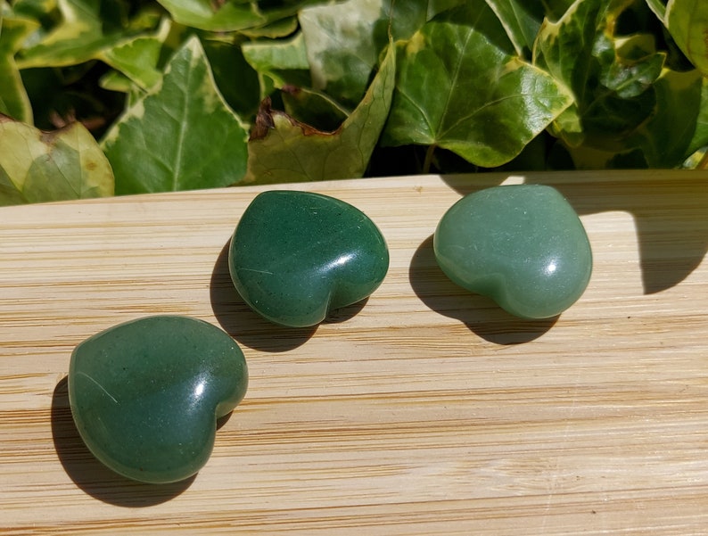 Pu&ograve; includere: Tre pietre di avventurina verdi a forma di cuore su una superficie di legno. Le pietre variano in tonalit&agrave;, da un verde foresta intenso a un verde pi&ugrave; chiaro e pastello. Lo sfondo presenta un fogliame verde.