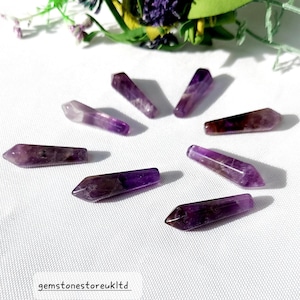 May include: Eight amethyst crystal wands arranged in a circular pattern. The wands are a deep purple colour with lighter purple and white streaks. The wands are pointed at one end and have a flat, polished surface. The text "gemstonestoreukltd" is at the bottom.