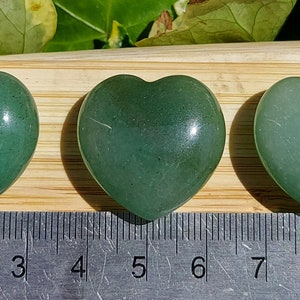 Pu&ograve; includere: Tre pietre a forma di cuore in avventurina verde, di diverse tonalit&agrave;, disposte in fila. Un righello con segni in centimetri &egrave; posto sotto. Le pietre sono su una superficie di legno, con fogliame verde sullo sfondo.