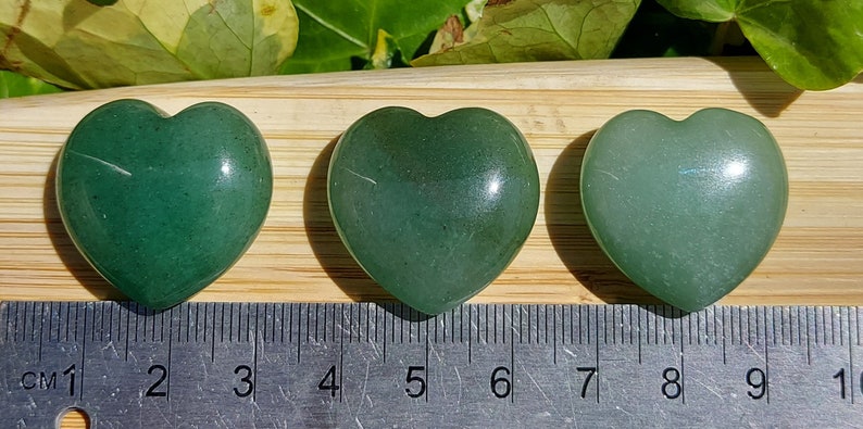 Pu&ograve; includere: Tre pietre a forma di cuore in avventurina verde, di diverse tonalit&agrave;, disposte in fila. Un righello con segni in centimetri &egrave; posto sotto. Le pietre sono su una superficie di legno, con fogliame verde sullo sfondo.