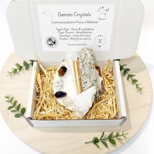 Może przedstawiać: Białe pudełko z napisem "Gemini Crystals" zawiera różne kryształy, wiązkę szałwii i patyczki do kadzideł. Pudełko zawiera kryształy Tygrysiego Oka, Kwarcu Przezroczystego, Ametystu i Howlitu. Pudełko stoi na drewnianej powierzchni z dekoracyjną zielenią.