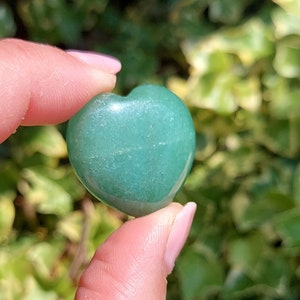Pu&ograve; includere: Una pietra preziosa verde lucidata a forma di cuore. La pietra &egrave; tenuta tra le dita, con uno sfondo sfocato verde e giallo di fogliame. La pietra preziosa &egrave; di colore verde opaco.