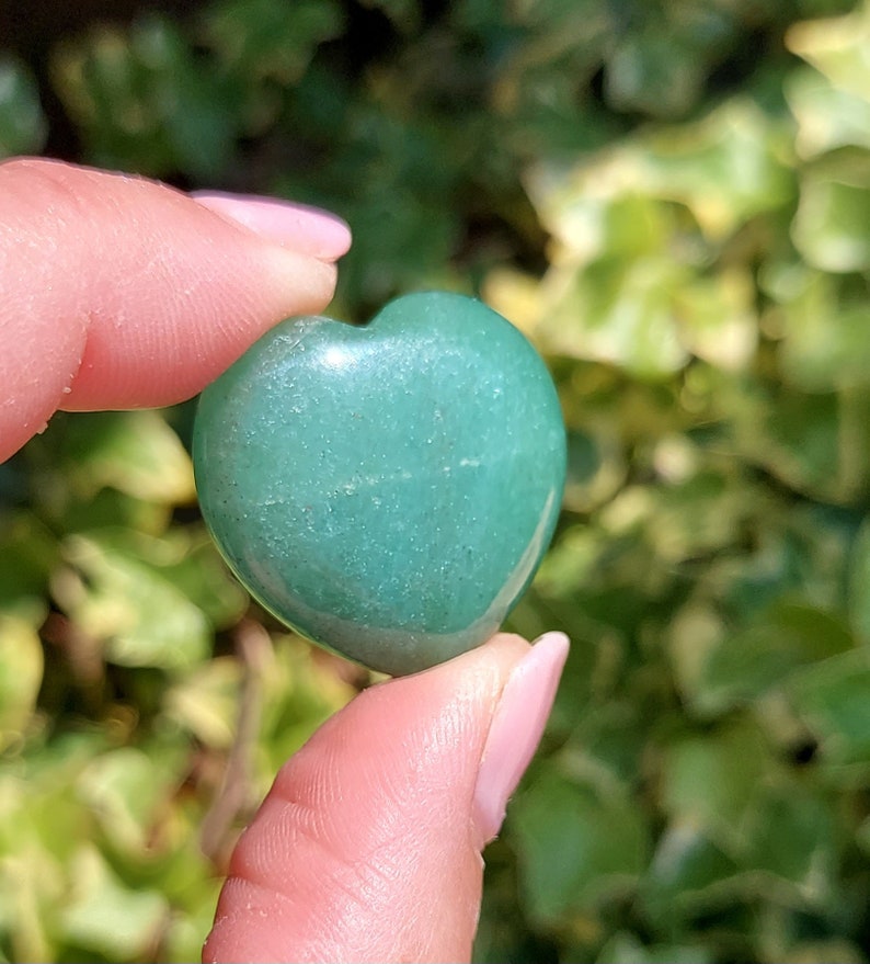 Pu&ograve; includere: Una pietra preziosa verde lucidata a forma di cuore. La pietra &egrave; tenuta tra le dita, con uno sfondo sfocato verde e giallo di fogliame. La pietra preziosa &egrave; di colore verde opaco.