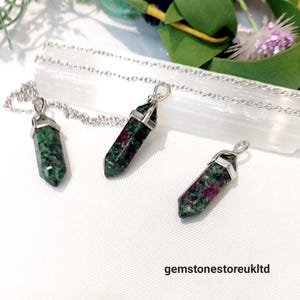 May include: Three green and pink gemstone pendants on silver chains. The pendants are in the shape of a point and are all different sizes. The pendants are on a white surface.