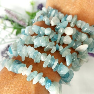 K&ouml;nnte beinhalten: Mehrere Str&auml;nge Aquamarin-Edelsteinarmb&auml;nder. Die Armb&auml;nder bestehen aus kleinen, unregelm&auml;&szlig;igen, hellblauen und wei&szlig;en Steinen. Die Armb&auml;nder sind um ein Handgelenk gewickelt, mit einem weichen, verschwommenen Hintergrund.