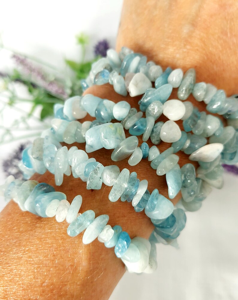 K&ouml;nnte beinhalten: Mehrere Str&auml;nge Aquamarin-Edelsteinarmb&auml;nder. Die Armb&auml;nder bestehen aus kleinen, unregelm&auml;&szlig;igen, hellblauen und wei&szlig;en Steinen. Die Armb&auml;nder sind um ein Handgelenk gewickelt, mit einem weichen, verschwommenen Hintergrund.