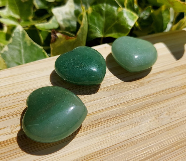 Pu&ograve; includere: Tre pietre verdi a forma di cuore su una superficie di legno. Le pietre hanno una consistenza lucida e liscia e variano leggermente nella tonalit&agrave;. Lo sfondo presenta un fogliame verde, aggiungendo un elemento naturale alla composizione.
