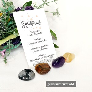 May include: A collection of gemstones for the Sagittarius zodiac sign. The image includes a card with the word "Sagittarius" and the names of the stones: Tiger's Eye, Amethyst, Snowflake Obsidian, and Citrine. The gemstones are in shades of brown, purple, and yellow.