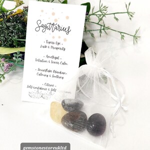 May include: A white card with the word "Sagittarius" and a list of gemstones: Tiger's Eye, Amethyst, Snowflake Obsidian, and Citrine. A small white organza bag contains several polished stones. The background includes greenery.