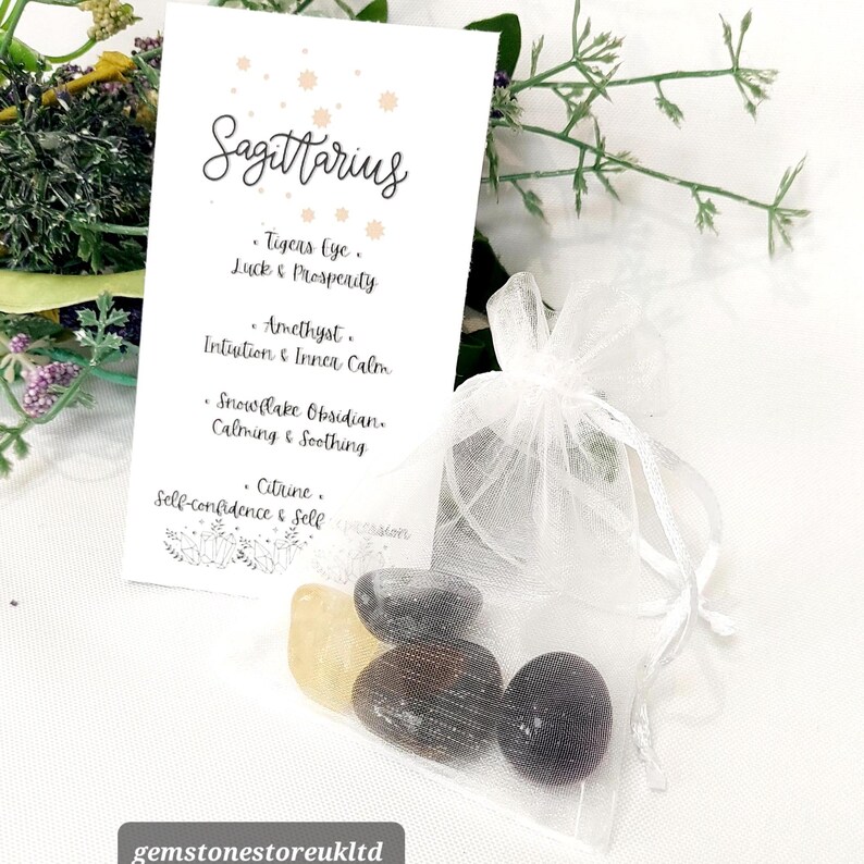 May include: A white card with the word "Sagittarius" and a list of gemstones: Tiger's Eye, Amethyst, Snowflake Obsidian, and Citrine. A small white organza bag contains several polished stones. The background includes greenery.