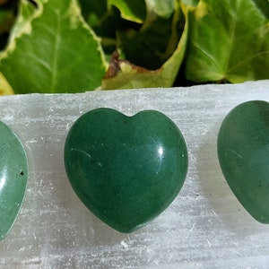 Pu&ograve; includere: Tre pietre verdi a forma di cuore, di diverse tonalit&agrave;, disposte in fila. Le pietre sono lisce e lucide. Lo sfondo presenta una superficie bianca e fogliame verde.