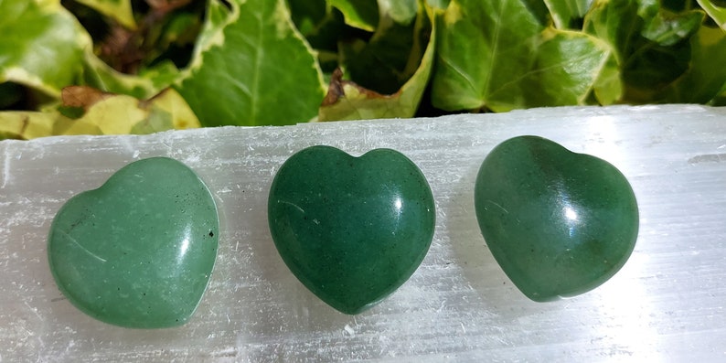 Pu&ograve; includere: Tre pietre verdi a forma di cuore, di diverse tonalit&agrave;, disposte in fila. Le pietre sono lisce e lucide. Lo sfondo presenta una superficie bianca e fogliame verde.