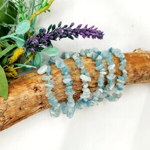 K&ouml;nnte beinhalten: Mehrere Armb&auml;nder aus Aquamarin-Chip-Perlen werden auf einem St&uuml;ck Treibholz pr&auml;sentiert. Die Armb&auml;nder sind hellblau und sind aufgef&auml;delt. Der Hintergrund umfasst lila Lavendel und gr&uuml;nes Laub.