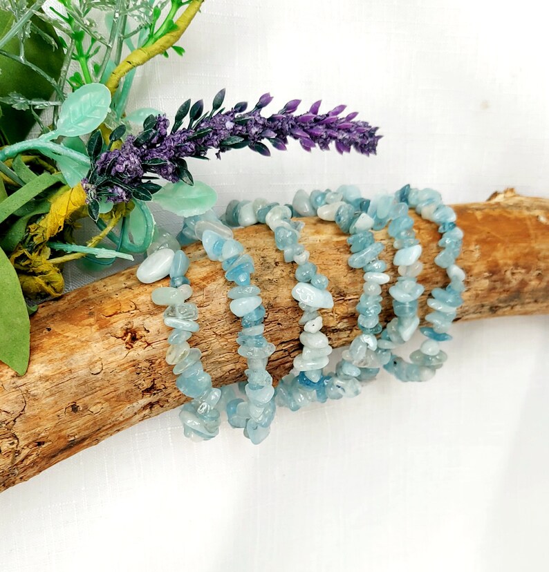K&ouml;nnte beinhalten: Mehrere Armb&auml;nder aus Aquamarin-Chip-Perlen werden auf einem St&uuml;ck Treibholz pr&auml;sentiert. Die Armb&auml;nder sind hellblau und sind aufgef&auml;delt. Der Hintergrund umfasst lila Lavendel und gr&uuml;nes Laub.