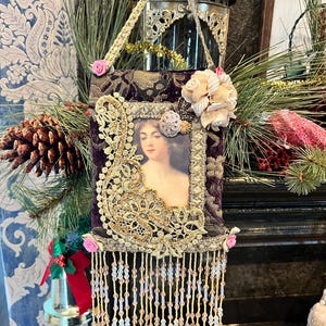 May include: A decorative ornament featuring a portrait of a woman, framed in a dark purple velvet fabric with gold lace and bead embellishments. The ornament is adorned with small pink roses and a beaded fringe, and is suspended by a gold ribbon.