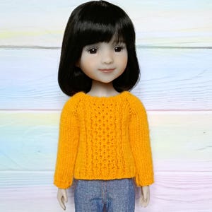 May include: A doll with black hair wearing a bright yellow knitted sweater and blue jeans. The sweater has a textured pattern on the front and long sleeves. The doll has a fair complexion and is smiling.