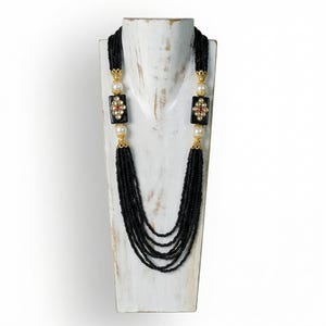 Puede incluir: Un collar largo de cuentas negras con detalles dorados, perlas y colgantes rectangulares negros con dise&ntilde;os florales y detalles rojos y dorados. El collar se exhibe en un busto de madera blanca.