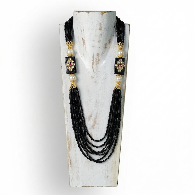 Puede incluir: Un collar largo de cuentas negras con detalles dorados, perlas y colgantes rectangulares negros con dise&ntilde;os florales y detalles rojos y dorados. El collar se exhibe en un busto de madera blanca.