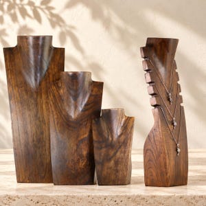 Könnte beinhalten: Ein Set aus vier dunkelbraunen Schmuckbüsten aus Holz in verschiedenen Höhen. Drei Büsten sind glatt und für Halsketten geformt, während die vierte an den Seiten Einkerbungen aufweist, an denen drei silberne Halsketten hängen.