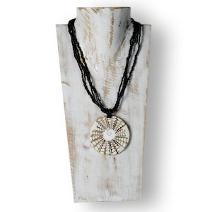 Puede incluir: Un collar artesanal con un colgante circular y hebras de cuentas negras. El colgante presenta una base color crema con un dise&ntilde;o geom&eacute;trico marr&oacute;n y una espiral blanca en el centro. El collar se exhibe en un expositor de joyas de madera blanca.