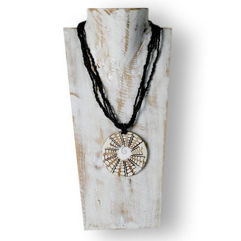 Puede incluir: Un collar artesanal con un colgante circular y hebras de cuentas negras. El colgante presenta una base color crema con un dise&ntilde;o geom&eacute;trico marr&oacute;n y una espiral blanca en el centro. El collar se exhibe en un expositor de joyas de madera blanca.