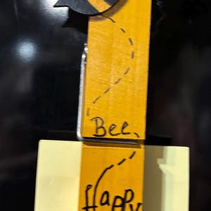 Könnte beinhalten: Eine holzgeschnitzte Bienenklammer mit gelbem Körper und schwarzen Streifen sowie weißen Flügeln. Die Klammer ist an einem gelben Haftzettel mit den Worten "Bee, Happy" in Schwarz befestigt.
