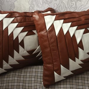 Op de afbeelding: Twee vierkante decoratieve kussens met een bruin leren buitenkant en witte driehoekige accenten. De kussens hebben een geometrisch ontwerp en liggen op een geruit oppervlak.