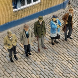 May include: A collection of miniature figurines depicting elderly people in period clothing, positioned on a cobblestone street. The figures include a man with a walking stick, a woman in a blue coat, and others in coats and hats, set against a building.