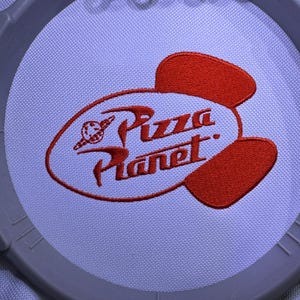May include: Embroidered Pizza Planet logo in red thread on white fabric, held in a gray embroidery hoop. The logo features the text "Pizza Planet" in a stylized font, with a planet and rocket ship graphic.
