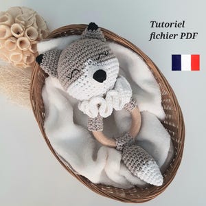 Może przedstawiać: Pluszowa zabawka w kształcie lisa z grzechotką, wykonana na szydełku w kolorze szarym i białym, z drewnianym pierścieniem. Zabawka znajduje się w wiklinowym koszyku wyłożonym białym kocem.