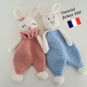 Peut inclure: Deux doudous en crochet en forme de lapin, l'un rose et l'autre bleu, avec des visages et des oreilles blancs. Les doudous portent des salopettes et ont un col à volants. Le texte "Tutoriel fichier PDF" est dans le coin supérieur droit de l'image.