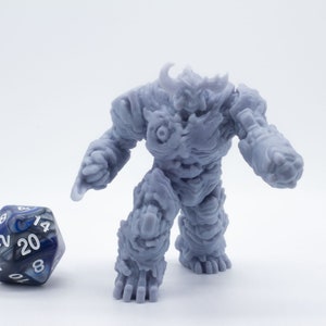 Puede incluir: Una miniatura gris y detallada de un golem de roca, una criatura mítica hecha de piedra, de pie sobre una superficie blanca. Un dado azul y blanco de 20 caras está en primer plano.
