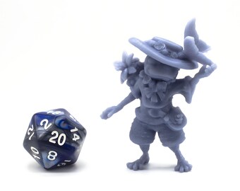 Op de afbeelding: Een blauwe en zilveren 20-zijdige dobbelsteen met witte nummers ligt naast een grijs 3D-geprint miniatuur van een kikker die een hoed draagt en een vis vasthoudt.