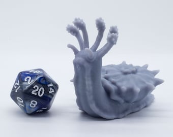 May include: A gray 3D printed miniature of a slug with five tentacles on its head. The slug is sitting on a white surface next to a blue and silver 20-sided die.