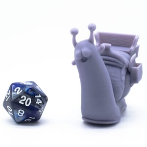 Puede incluir: Una figurita de caracol impresa en 3D de color morado con una cara sonriente y una pequeña silla de montar en la espalda. Un dado de 20 caras azul y plateado está en primer plano.