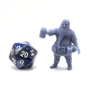 Puede incluir: Una miniatura azul grisáceo de un enano sosteniendo una jarra, junto a un dado de veinte caras. El dado es azul, negro y blanco. El enano tiene una larga barba y lleva un cinturón con bolsillos. Para juegos de mesa.