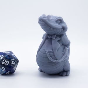 Könnte beinhalten: Eine graue 3D-gedruckte Figur eines Krokodils im Anzug mit Krawatte. Das Krokodil steht auf einer weißen Oberfläche neben einem blauen und silbernen 20-seitigen Würfel.