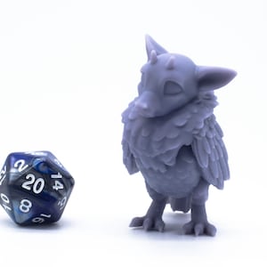 Op de afbeelding: Een paarse 3D-geprinte miniatuur van een klein, gevederd wezen met een lange snavel en grote ogen. Het wezen staat op een wit oppervlak naast een blauwe en zwarte 20-zijdige dobbelsteen.