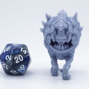 Puede incluir: Una miniatura gris de un monstruo impresa en 3D con una cabeza puntiaguda y dientes afilados. El monstruo está de pie sobre una superficie blanca junto a un dado de 20 caras azul y negro.