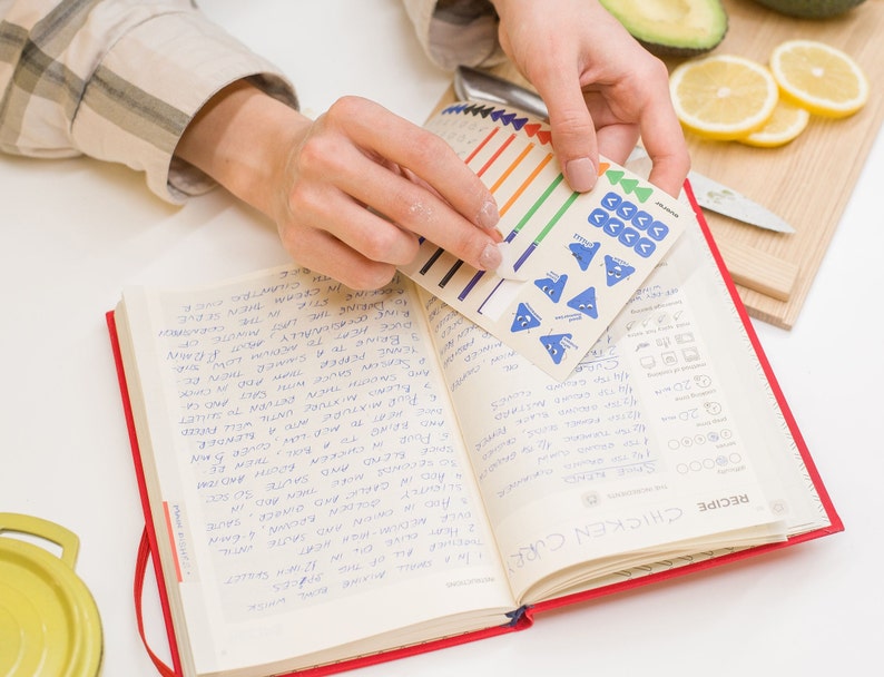 Puede incluir: Un libro de cocina rojo y blanco con notas escritas a mano y una hoja de pegatinas azul y blanca con flechas y s&iacute;mbolos. El libro de cocina est&aacute; abierto en una p&aacute;gina titulada "Curry de pollo" con una receta escrita a mano.